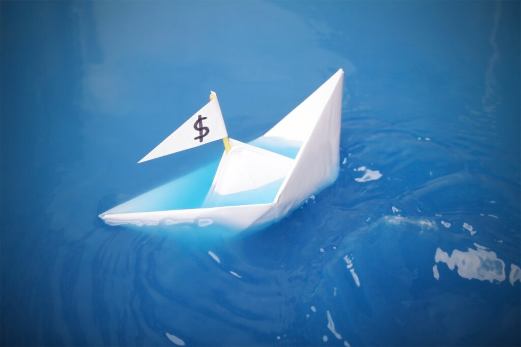 paper boat with a dollar sign on its flag is sinking in blue water