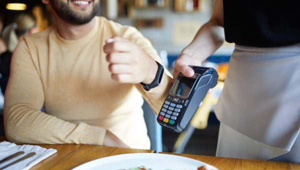 Smiling young man with smartwatch keeping his wrist close to payment machine while using transfer system to pay for meal